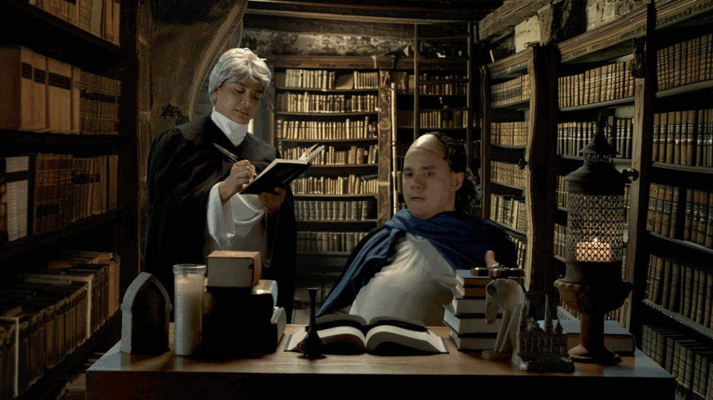 An image of a man sitting in front of a desk with another man standing to his left taking notes. In the background is a rustic library setting.