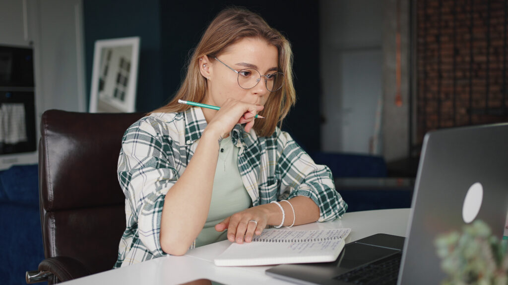 Still Image of Elearning Young Serious Woman Student Studying Online At Home Reading Information On Laptop Screen And Making Notes